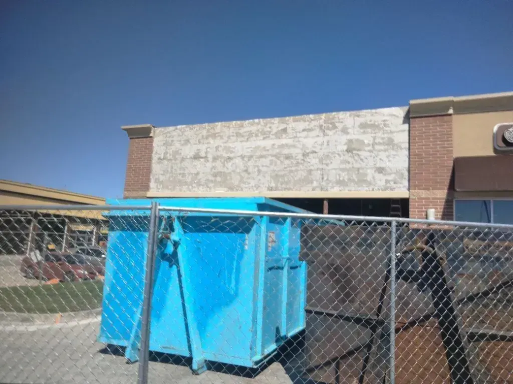 A bright blue dumpster sits in a parking lot in front of a building with a large, weathered, textured sign area.