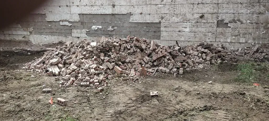 A pile of brick and concrete rubble sits on a patch of dirt in front of a weathered masonry wall.