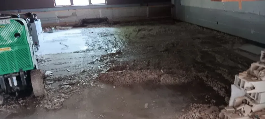 A green mini-excavator sits on a construction site floor covered in loose dirt and debris near a stack of concrete blocks.