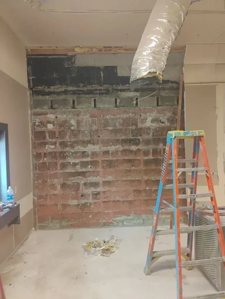 A construction site showing a brick wall with exposed framing and a duct overhead, next to an open stepladder.