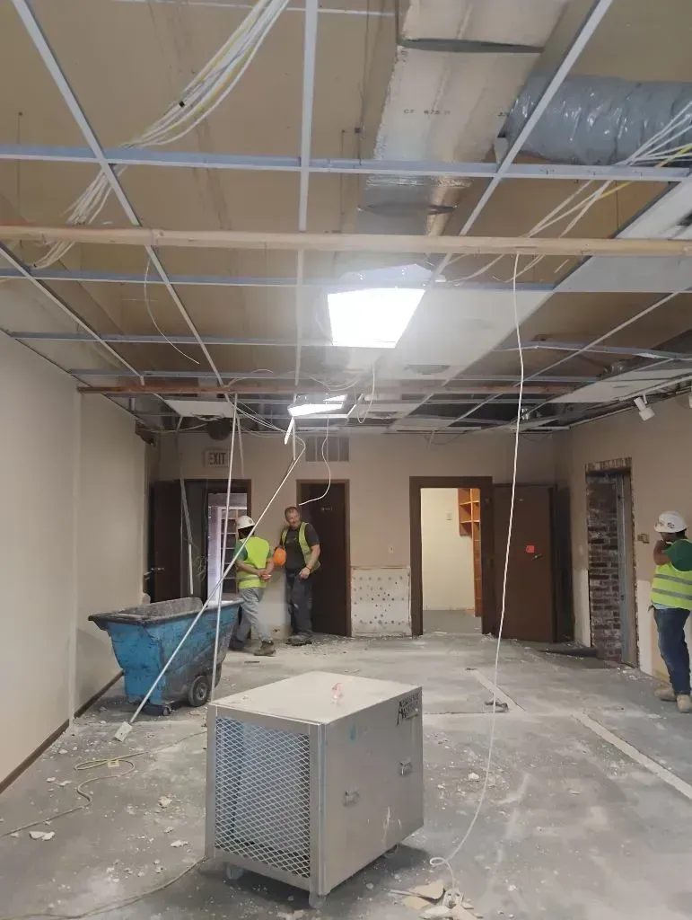 Three workers in high-visibility vests renovate an office with exposed ceiling grids, a dumpster, and industrial fans.