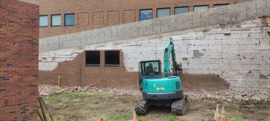 A teal excavator sits in front of a brick building with a partially exposed concrete foundation undergoing construction.