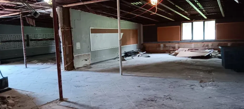 A large, dilapidated indoor space under renovation, featuring exposed ceiling beams, unfinished walls, and debris.
