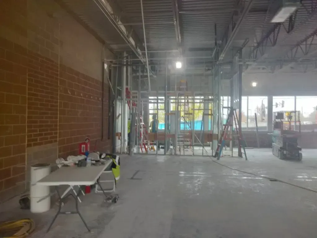 An indoor construction site features a folding table in the foreground, exposed metal framing, and a brick wall.