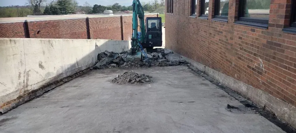 A teal excavator operates at the base of a concrete ramp between a brick building wall and a retaining wall.