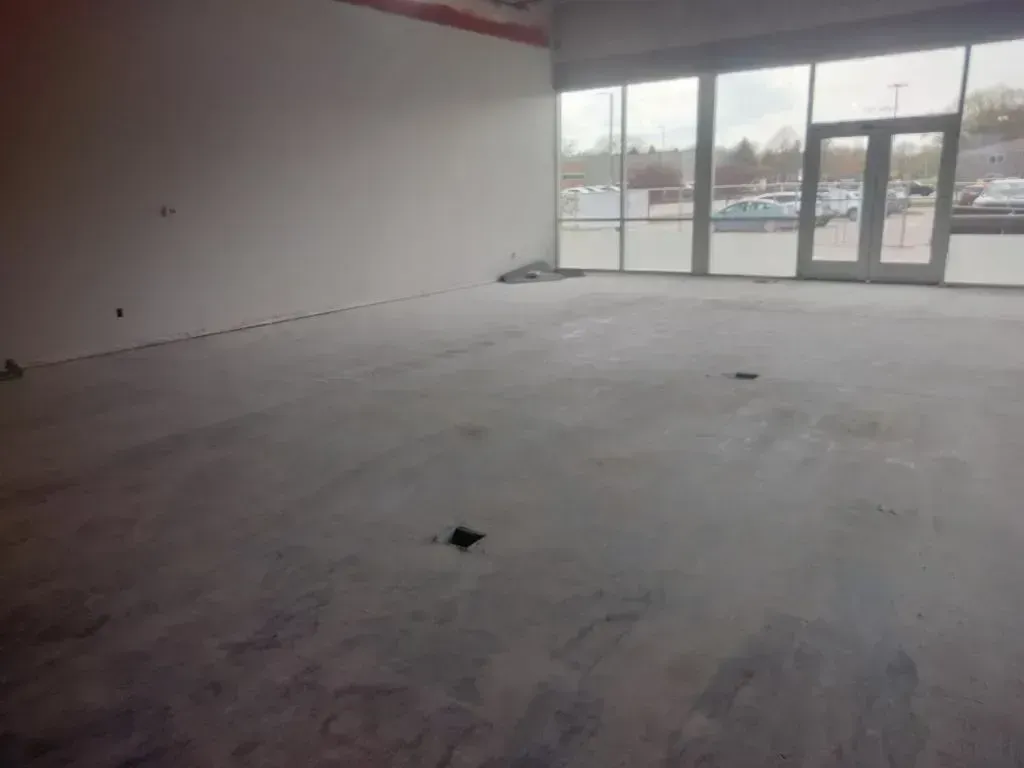 An empty, unfinished commercial interior with a concrete floor, white walls, and a large glass storefront entrance.
