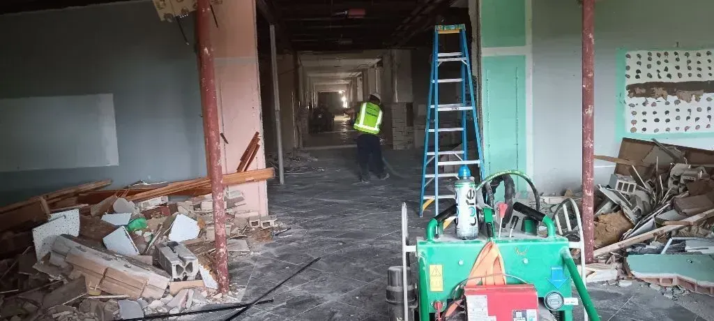 A construction worker in a high-visibility vest stands in a room undergoing interior demolition, near a blue ladder.