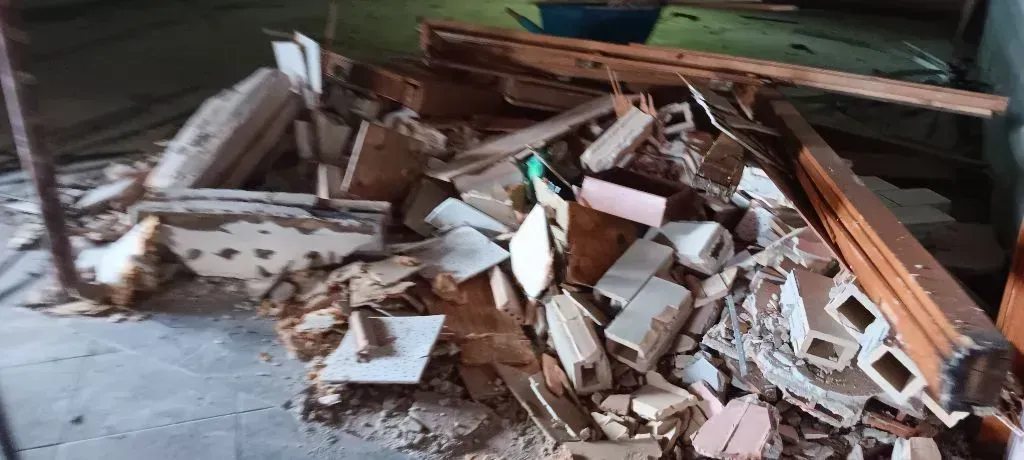 A pile of construction debris, including shattered white tiles, broken wooden beams, and masonry, on a concrete floor.