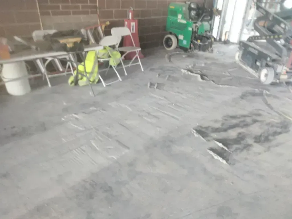 A view of a workshop floor with folding chairs, a green piece of machinery, and structural damage on the concrete surface.