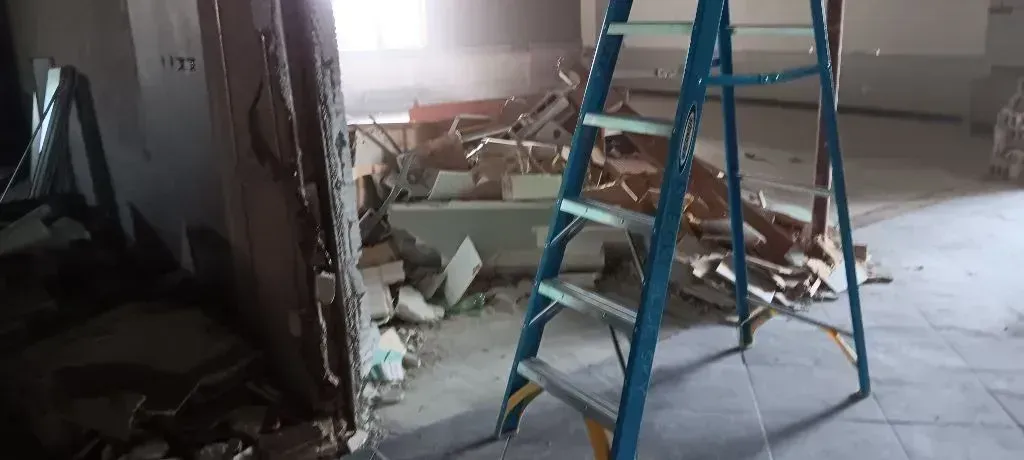 A bright blue stepladder stands in an indoor space under renovation, surrounded by construction debris and rubble.