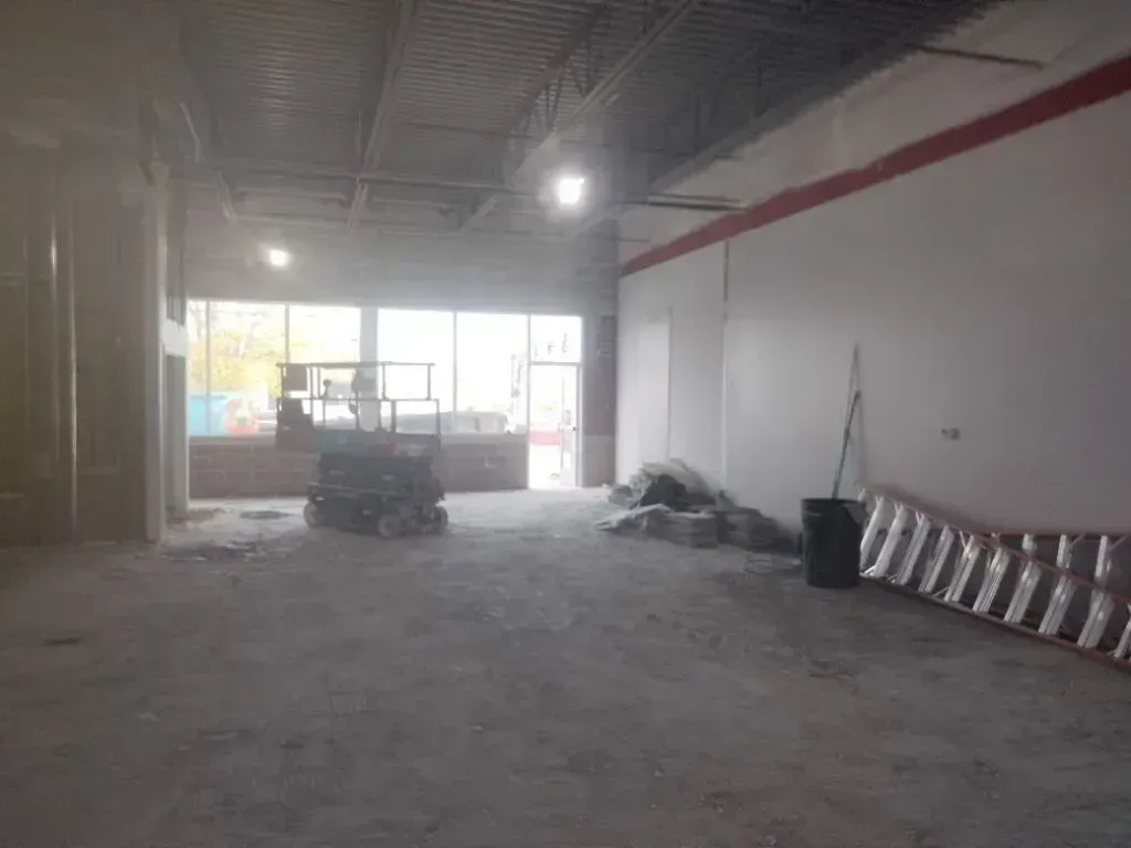 Empty, unfinished commercial space with exposed ceiling, concrete floors, large windows, and construction equipment.