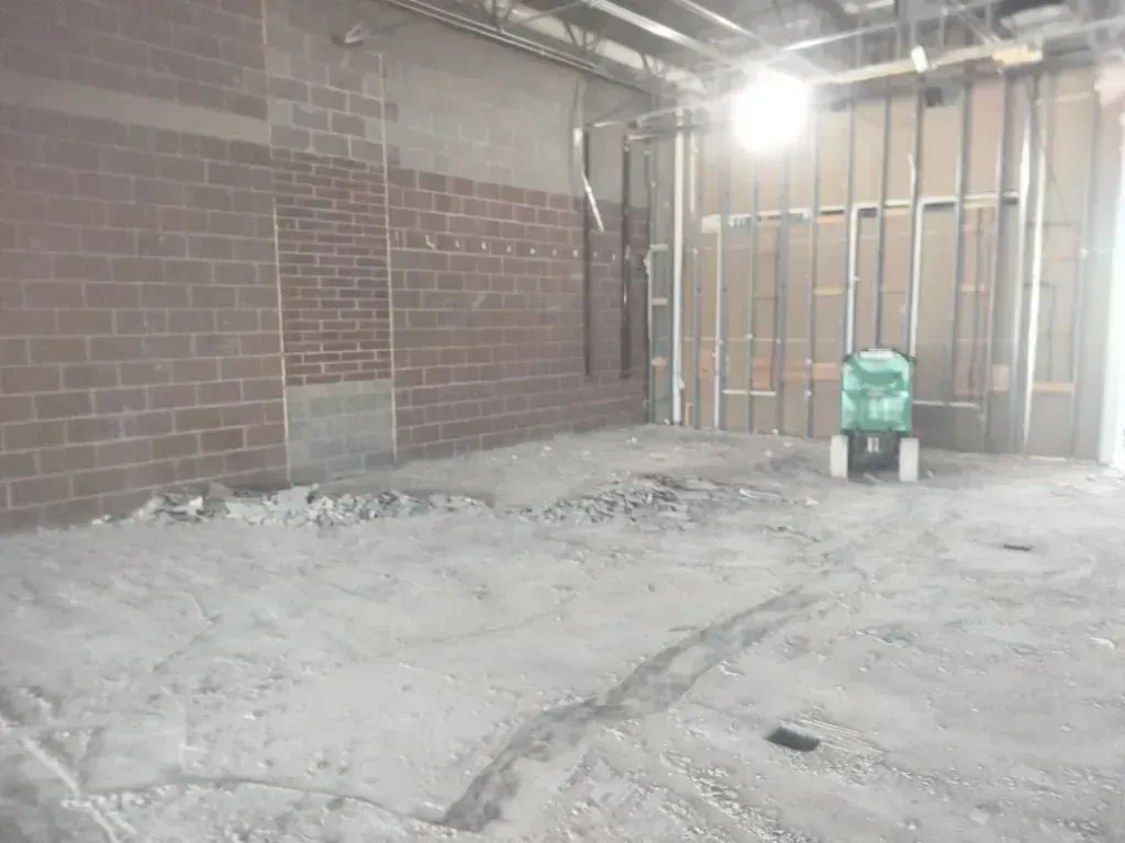 A construction site with a red brick wall, exposed metal framing, and a dusty concrete floor with debris.