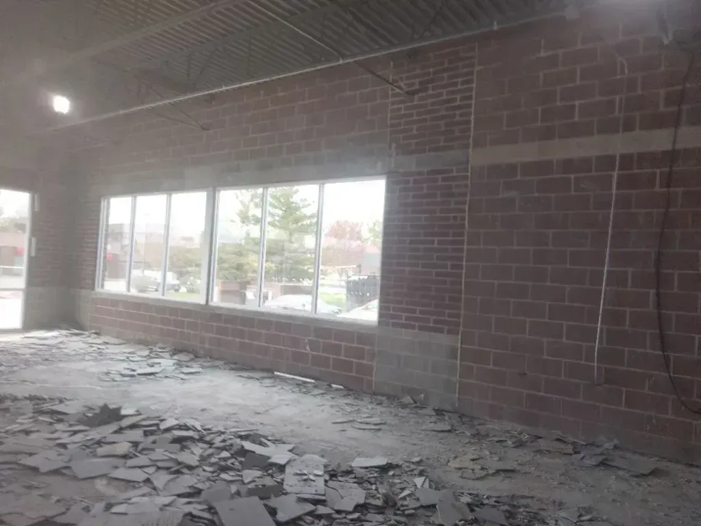 A construction site showing a brick wall with a large window opening and floor covered in scattered debris.