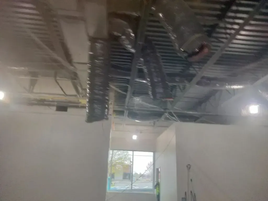 Construction view of an unfinished room interior showing exposed HVAC ductwork on a metal ceiling and a bright window.