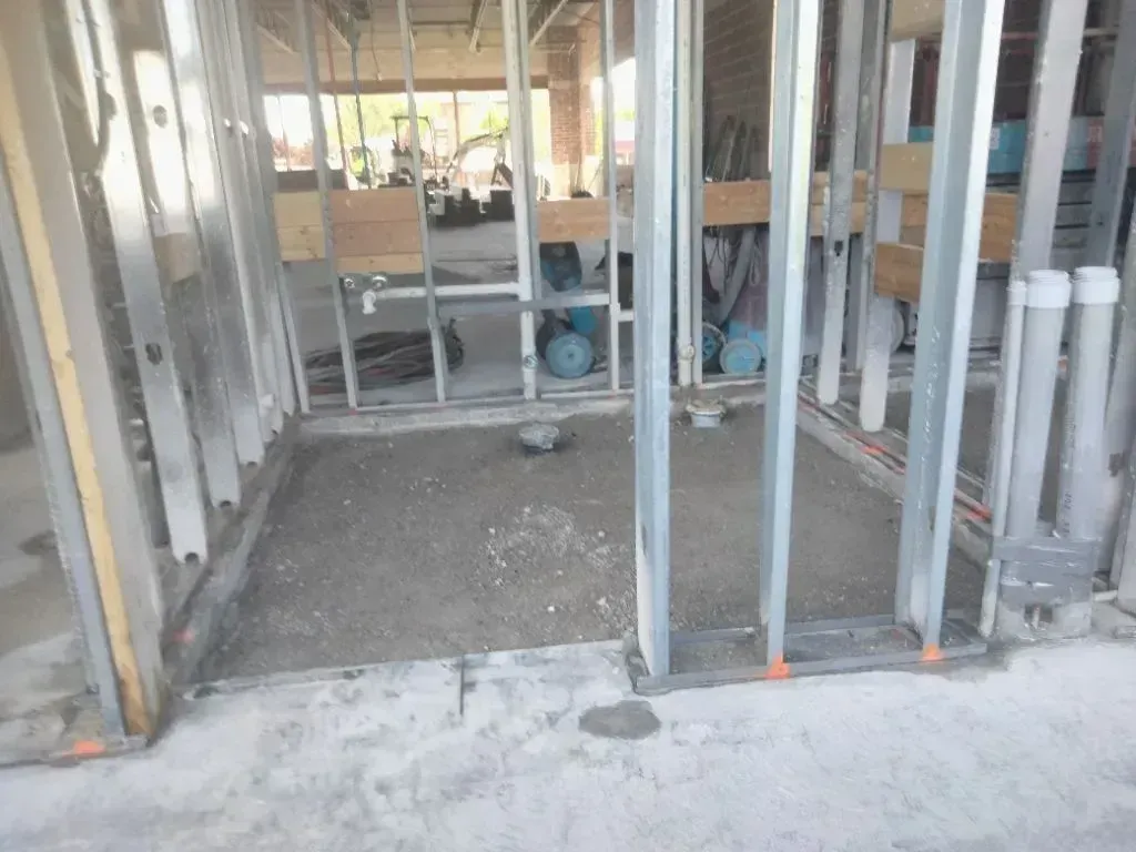 Construction site showing a framed wall opening for a bathroom area with a concrete floor and exposed plumbing pipes.