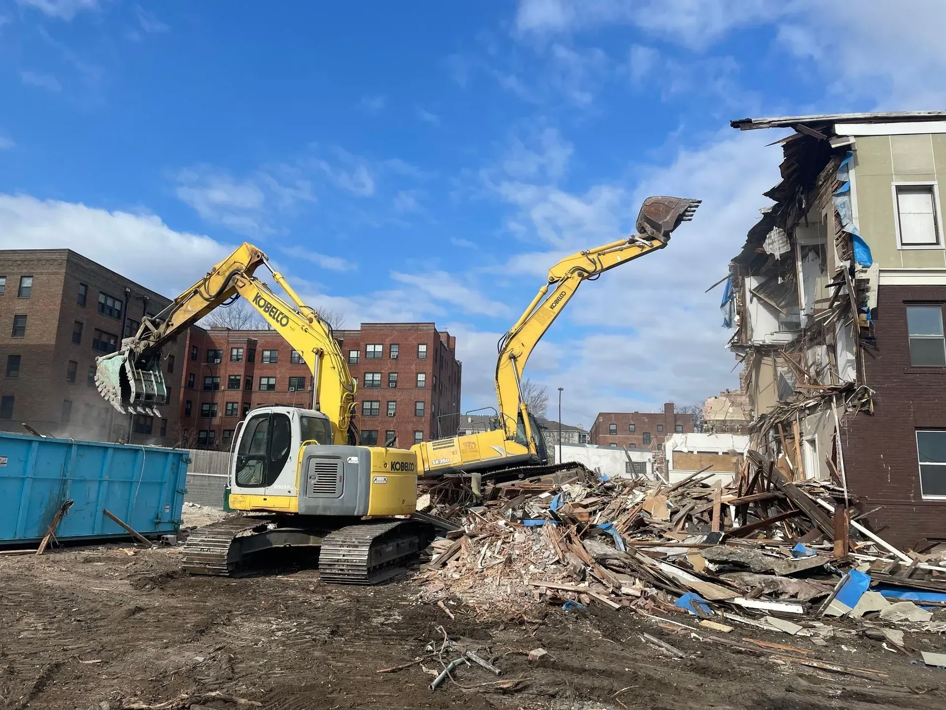 A worker in an orange shirt and hard hat sweeps construction debris into a trash bin in a room with gray block walls.