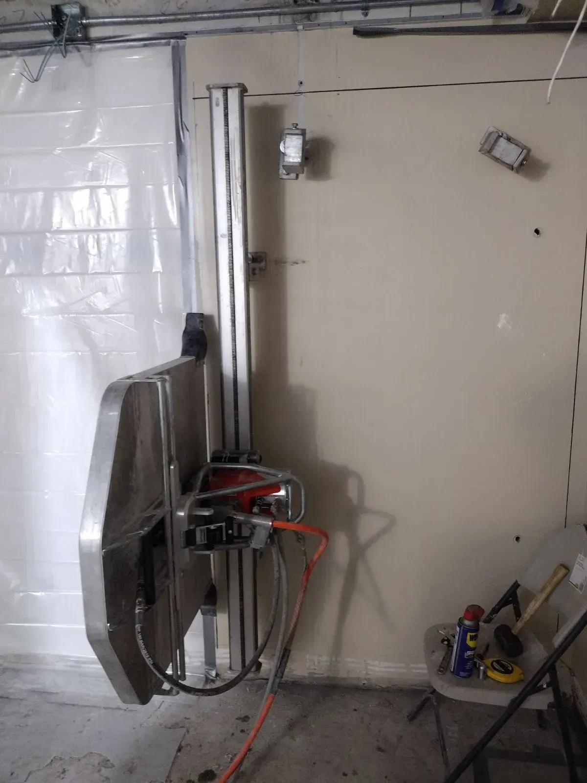 A wall saw mounted on a vertical track against an interior wall, with a chair and tools on the floor nearby.