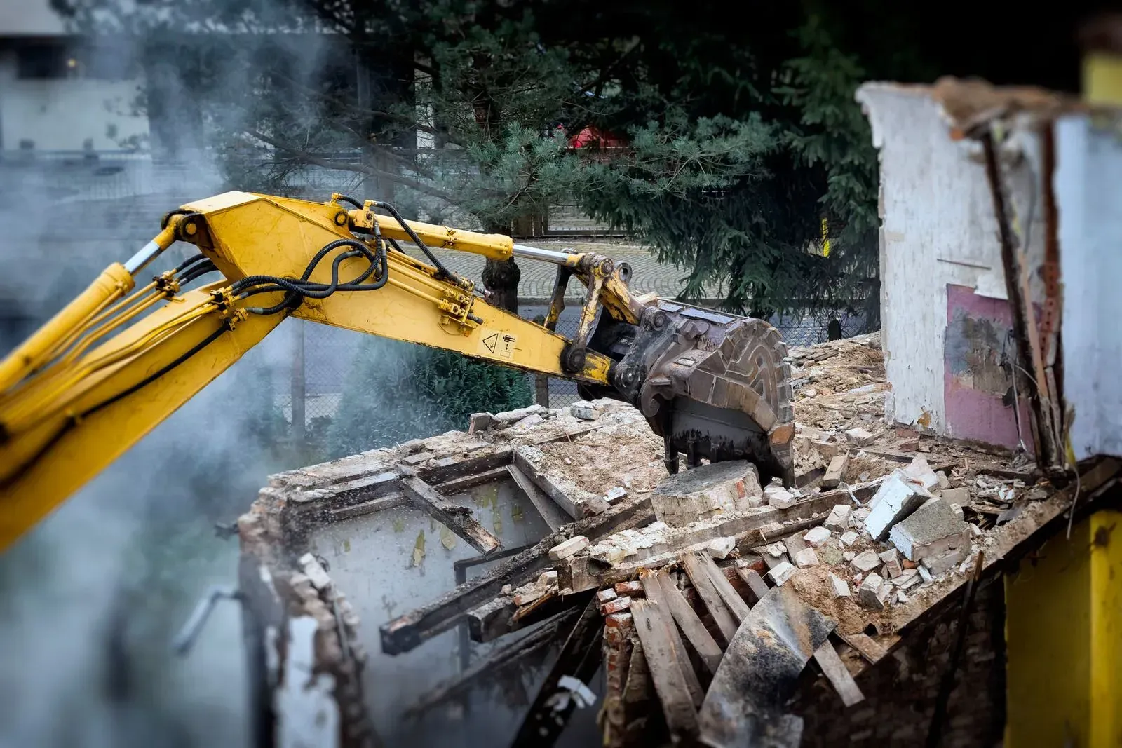 A yellow excavator arm demolishing the top level of a building, causing dust and debris to scatter.