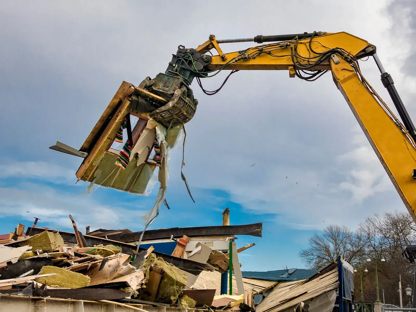 A yellow demolition excavator lifting debris from a pile of building rubble against a cloudy sky.