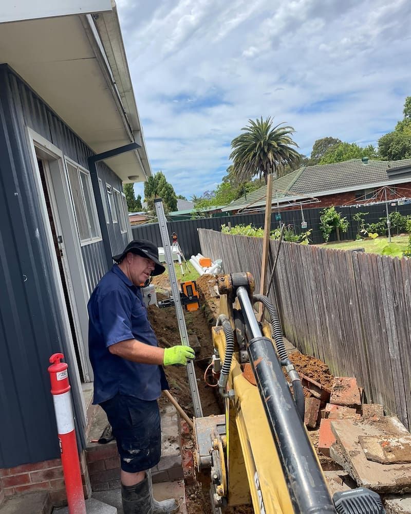 A Man is Standing Next to a Yellow Excavator in Front of a House — Bridge Plumbing & Trades in Nowra, NSW