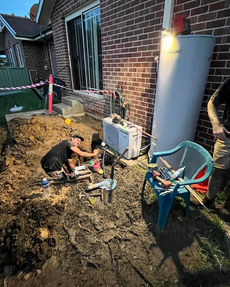 A Man is Working on a Water Heater Outside of a Brick House — Bridge Plumbing & Trades in Nowra, NSW