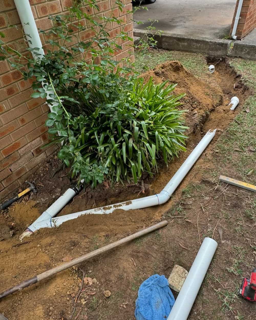 A Drain Pipe is Being Installed in a Garden Next to a Brick Wall — Bridge Plumbing & Trades in Nowra, NSW