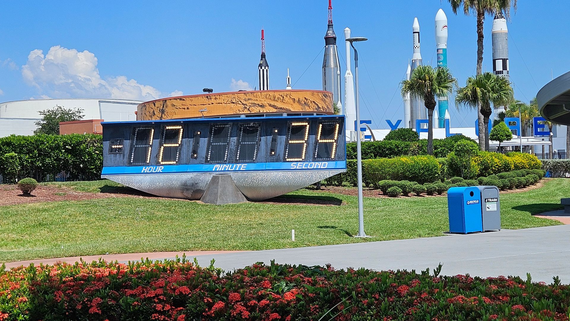 Digital clock, displaying numbers, on display at Kennedy Space Center with rockets in background.