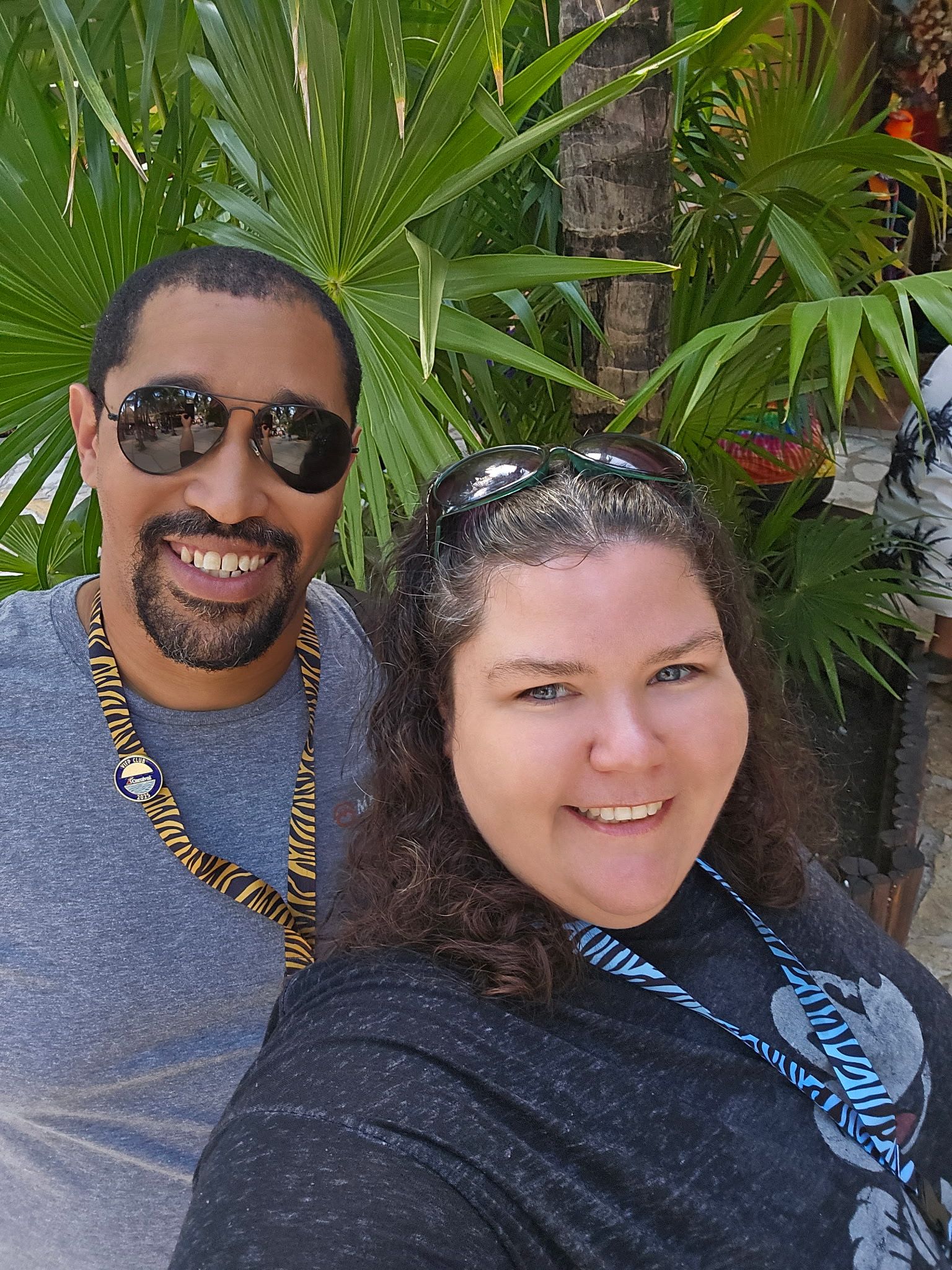Two people smiling, posing for a selfie in front of a tropical plant. One wears sunglasses and a gray shirt.