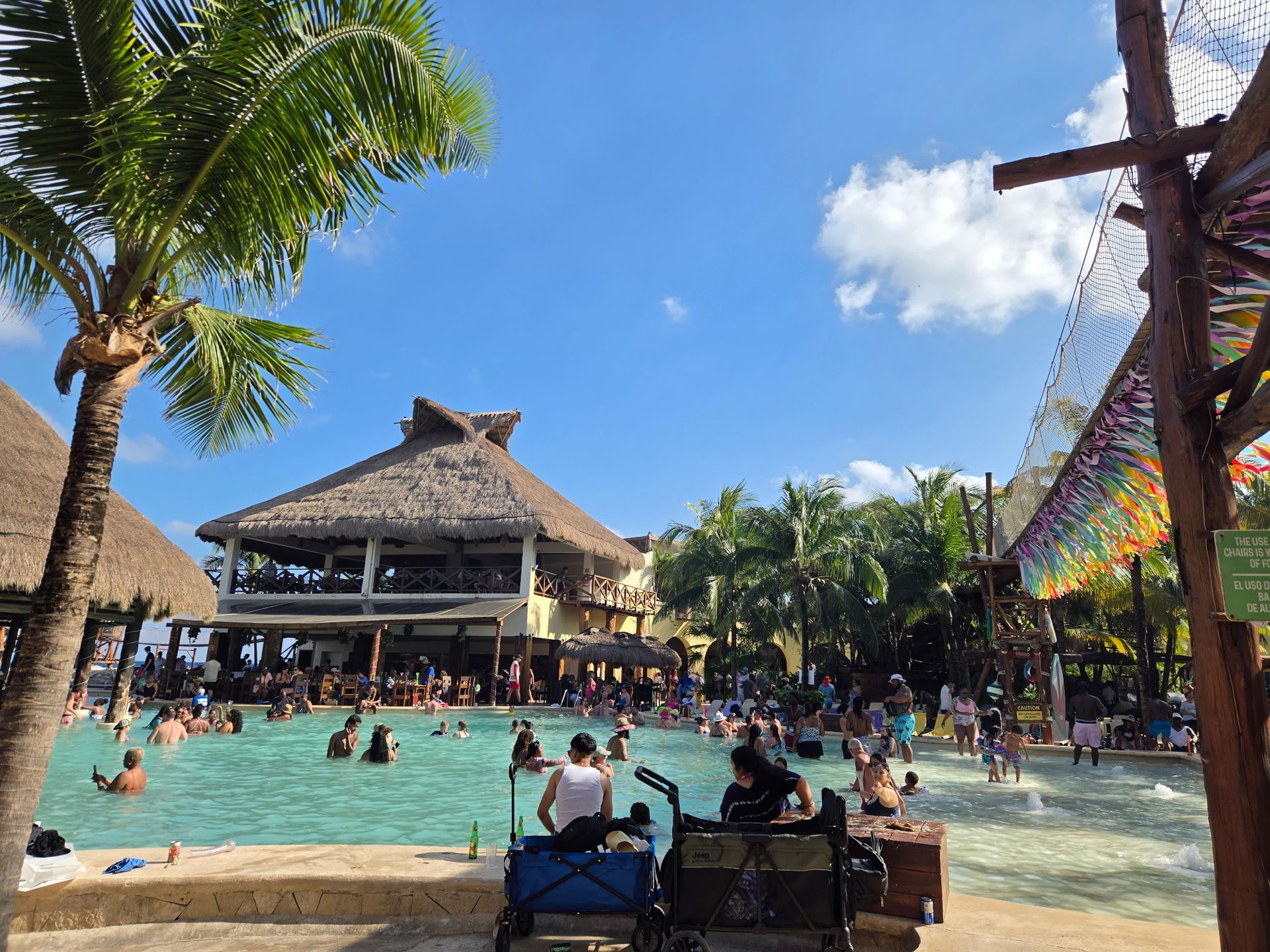 Poolside scene with many people swimming and enjoying drinks; thatched roof bar overhead. Blue sky.