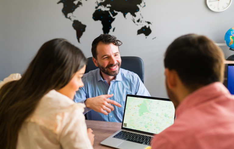 A smiling person in a blue shirt points at a laptop map while consulting with two others in an office.