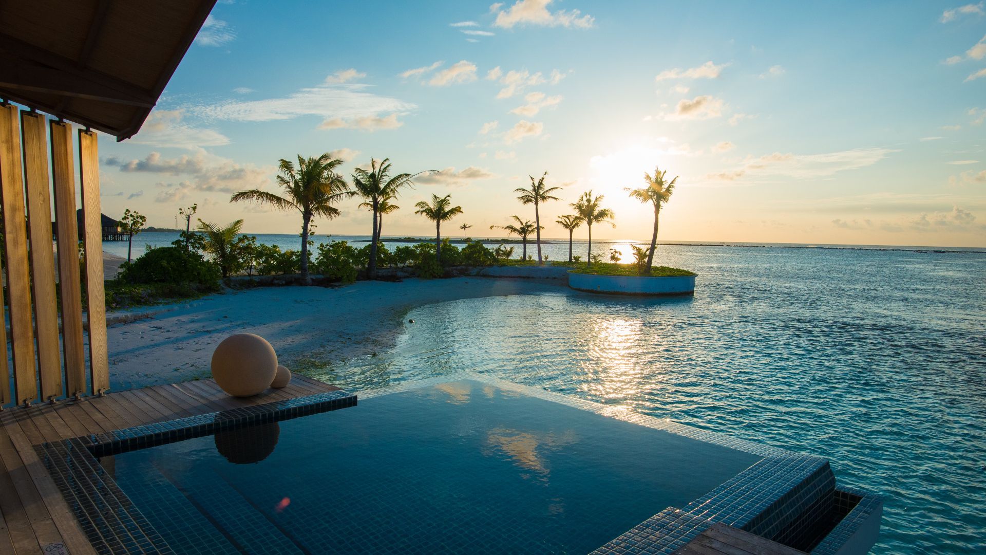 Infinity pool overlooking a tropical ocean at sunset, with palm trees and a resort patio.