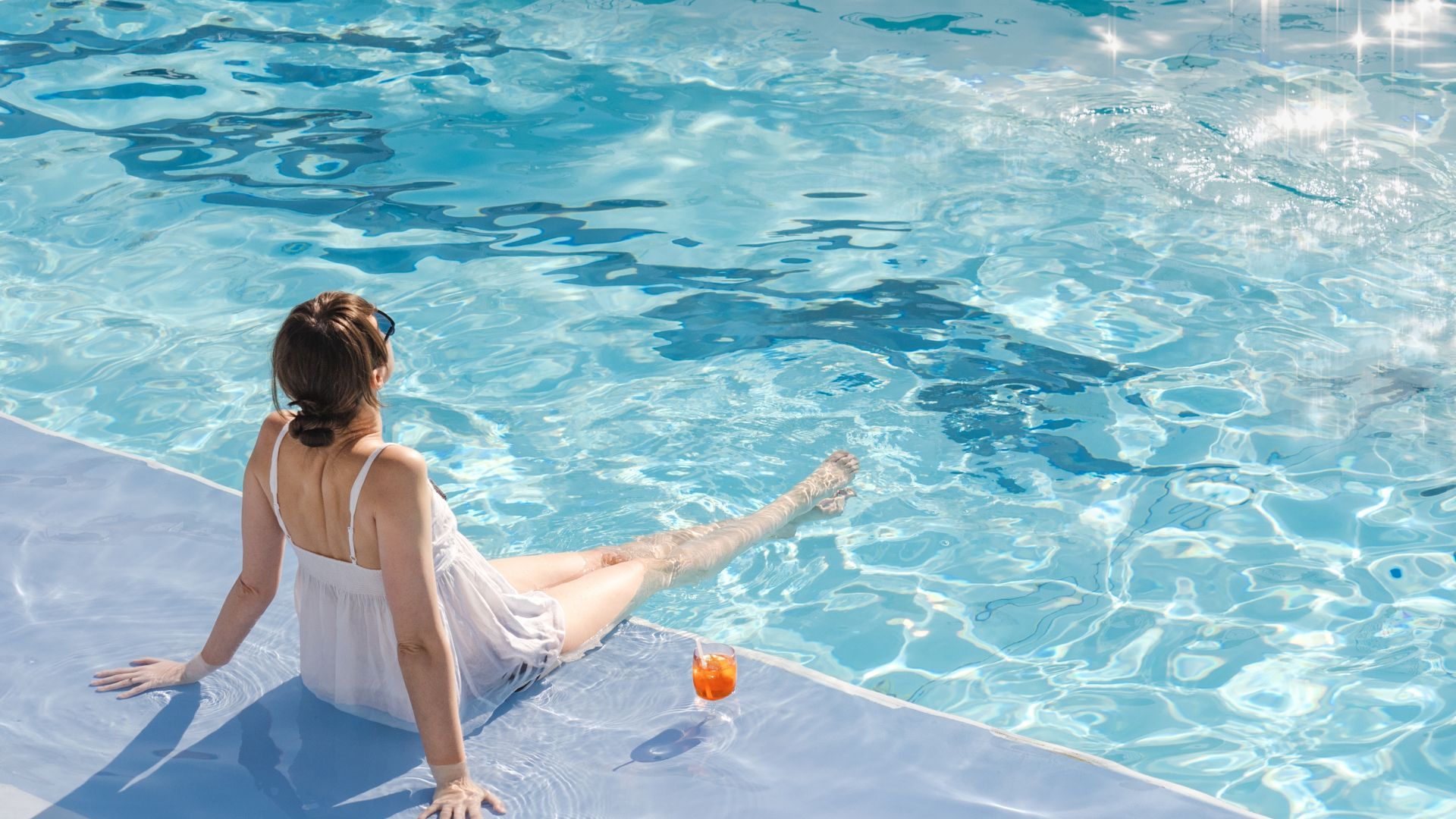 A woman is sitting on the edge of a swimming pool with a drink.
