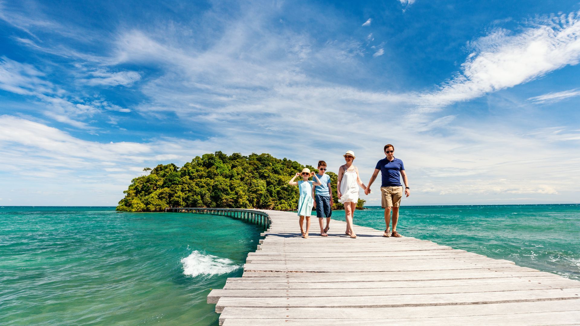 A family is walking across a wooden pier to a small island in the ocean.