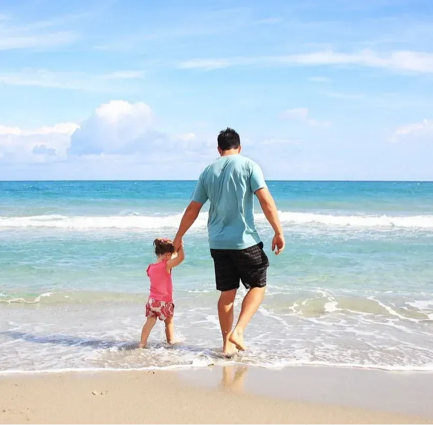 A man and a little girl are walking on the beach holding hands.