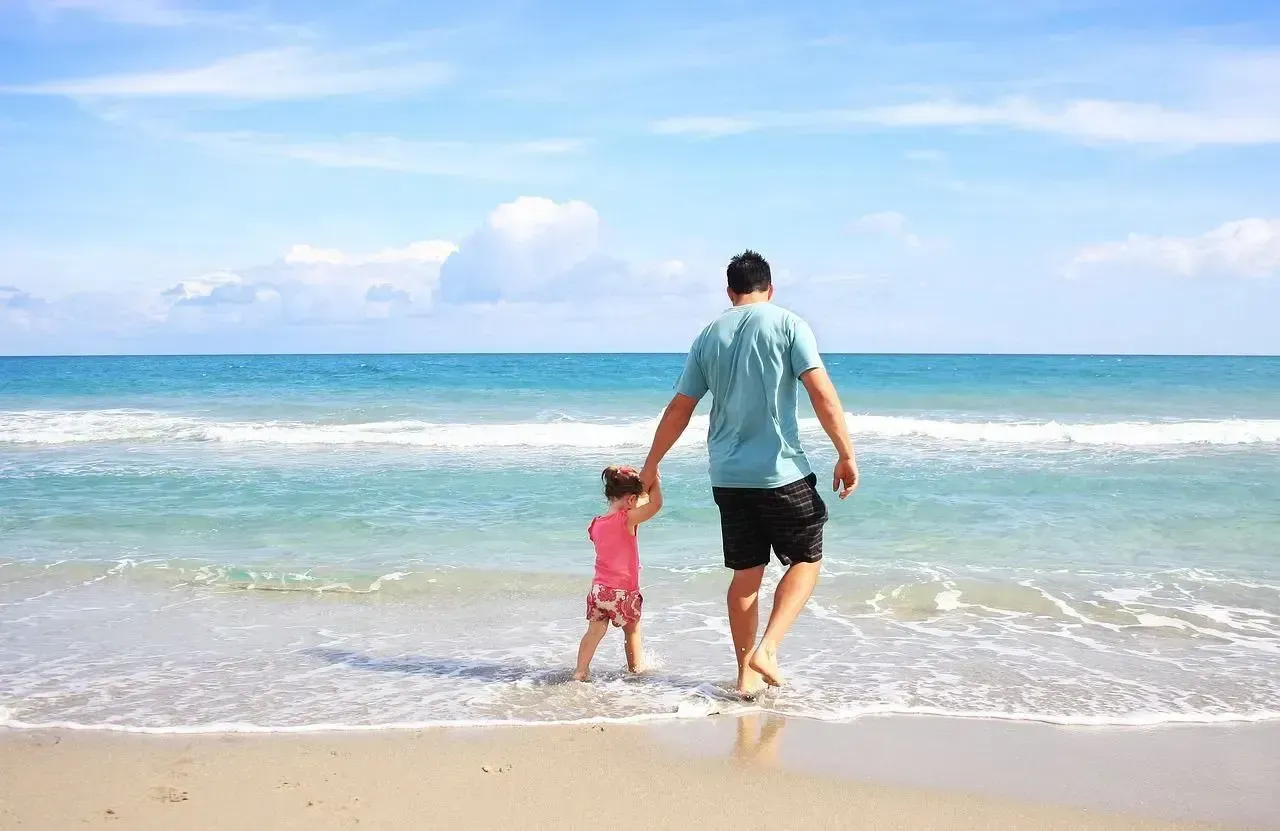 A man and a little girl are walking on the beach holding hands.