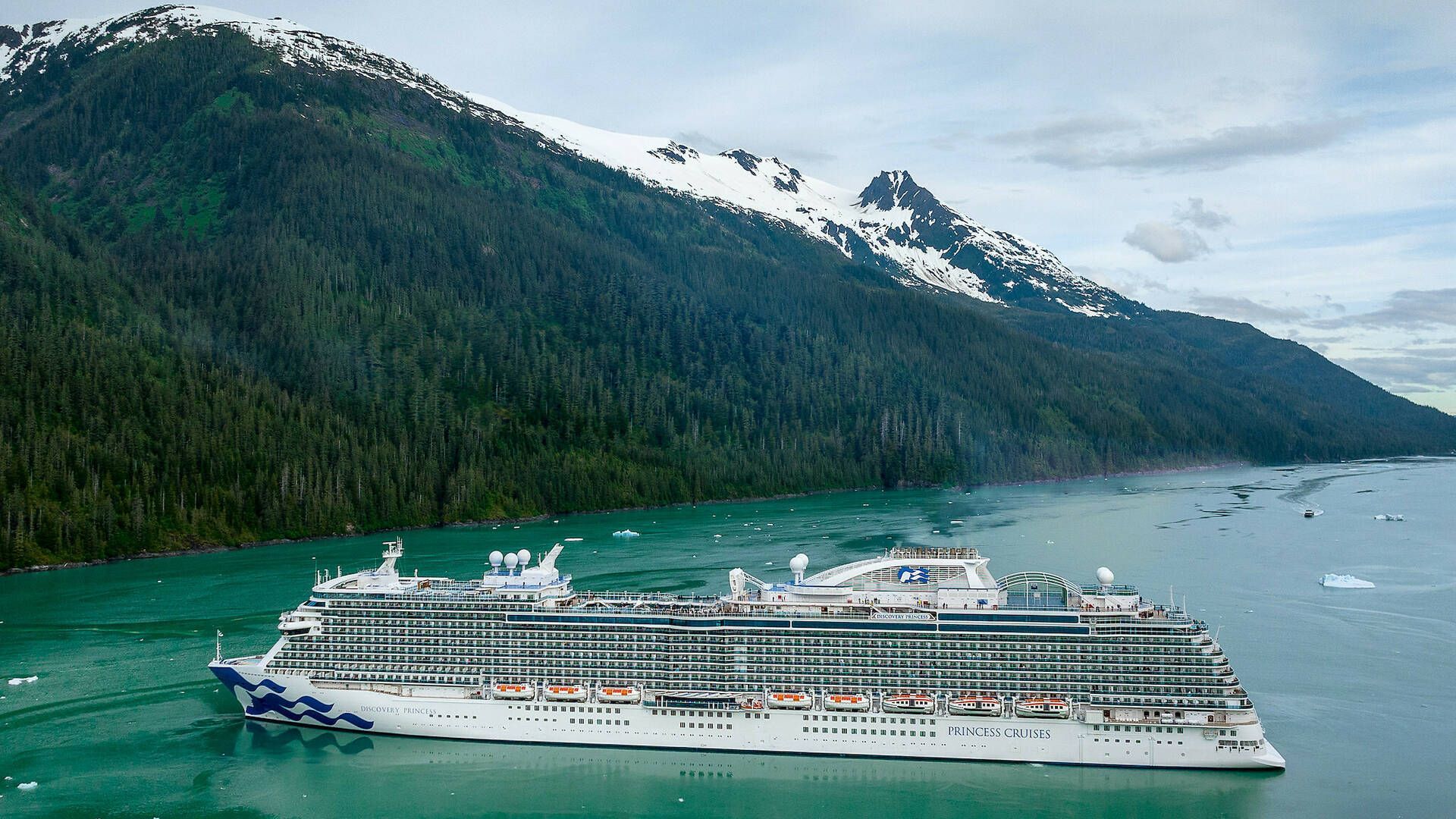 A large white cruise ship sails through turquoise water in front of a majestic, snow-capped mountain range and forest.