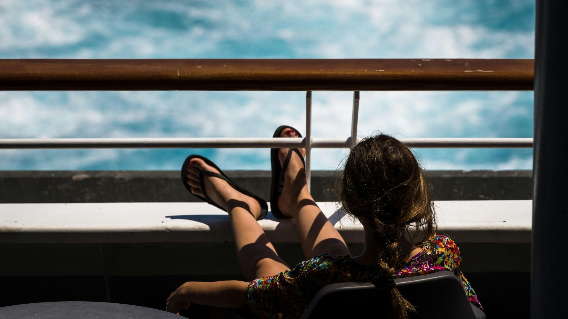 Person reclining on a deck chair, feet up, looking out at the ocean through a railing.