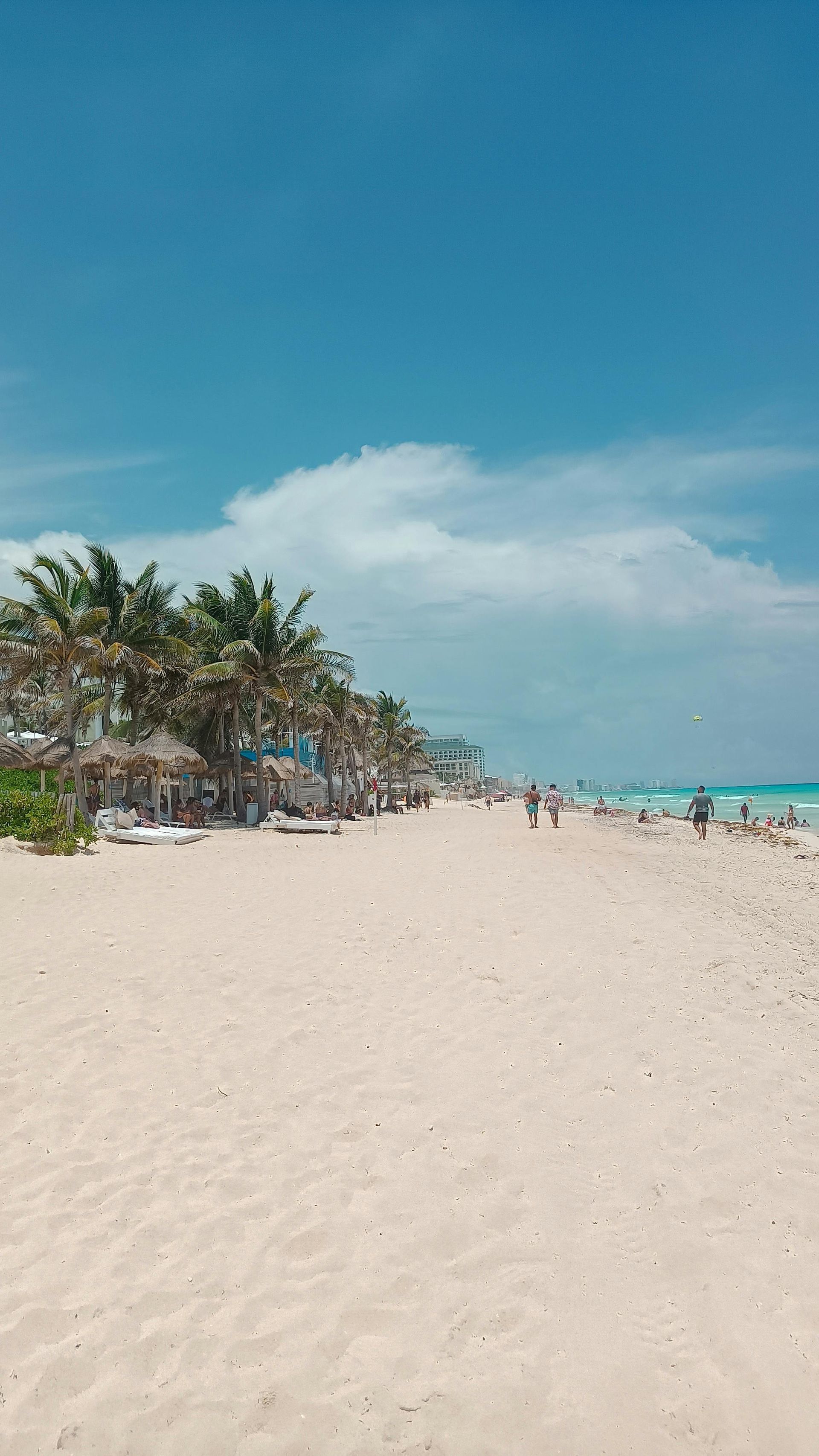 Sunny beach with white sand, turquoise water, palm trees, and a few distant people under a blue sky