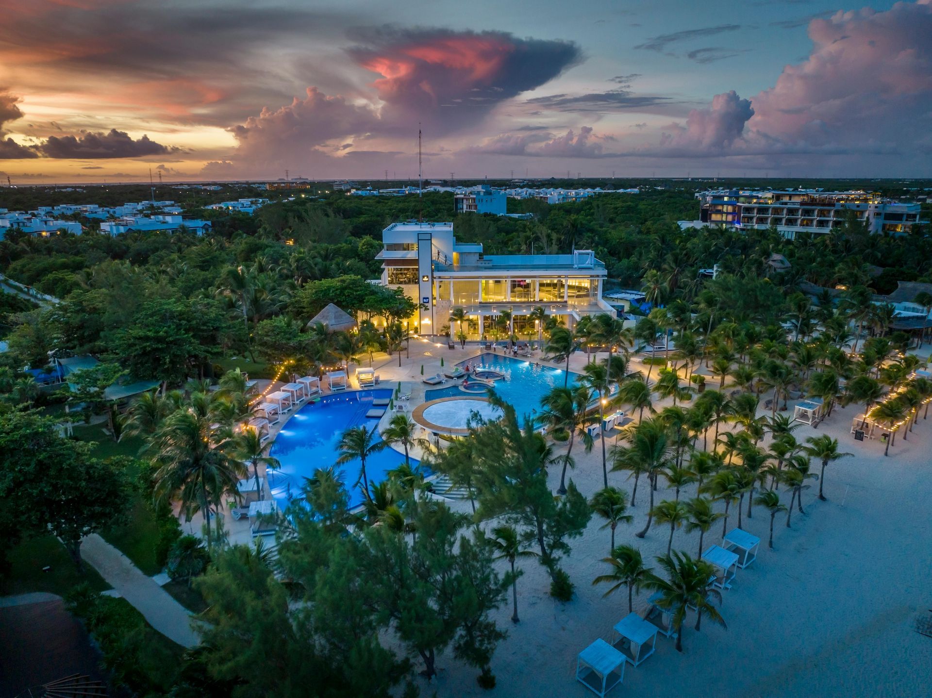Aerial view of a tropical resort with lit pool, palm trees, and sunset sky over the beach