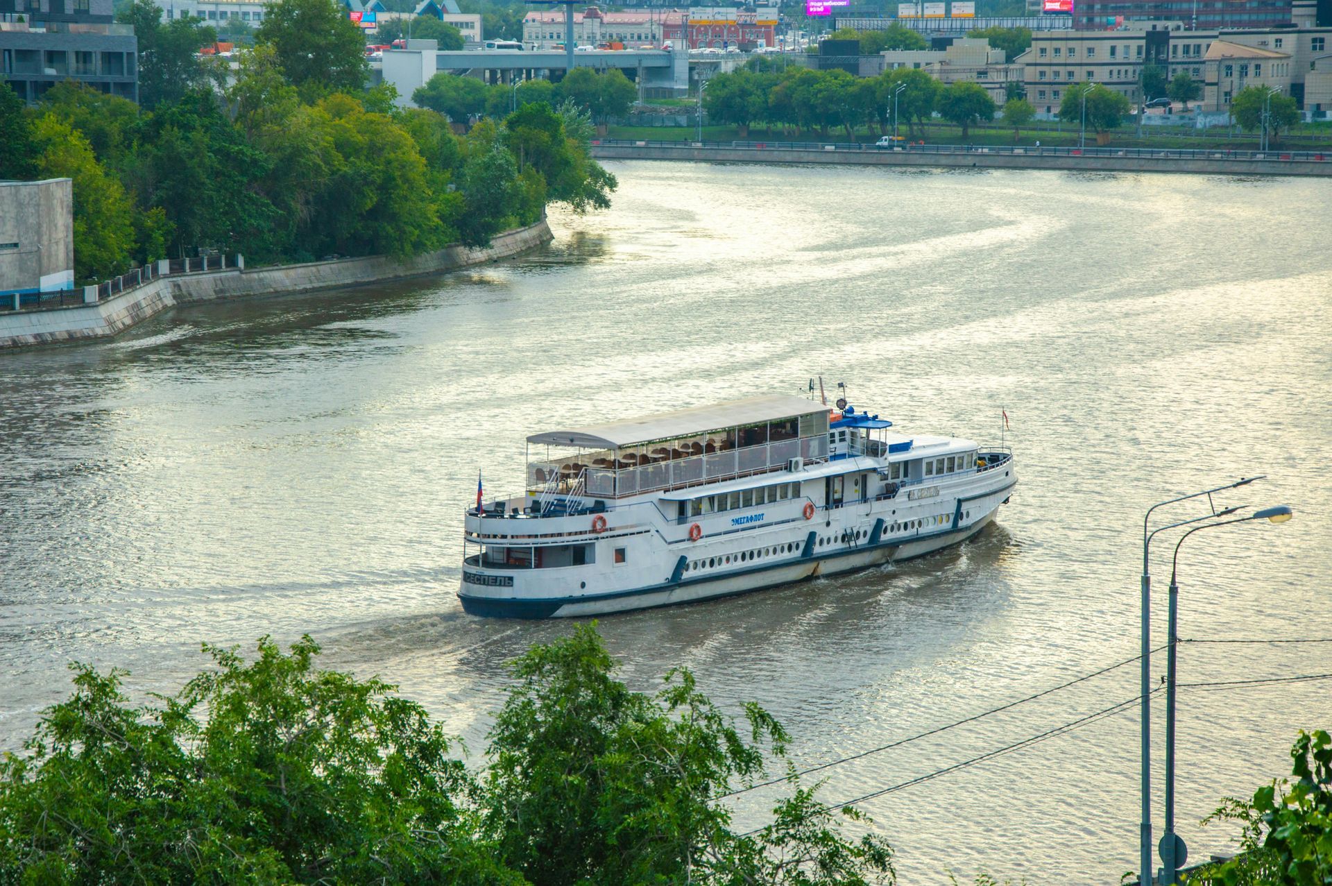 White passenger boat on a river near a green shoreline and city buildings
