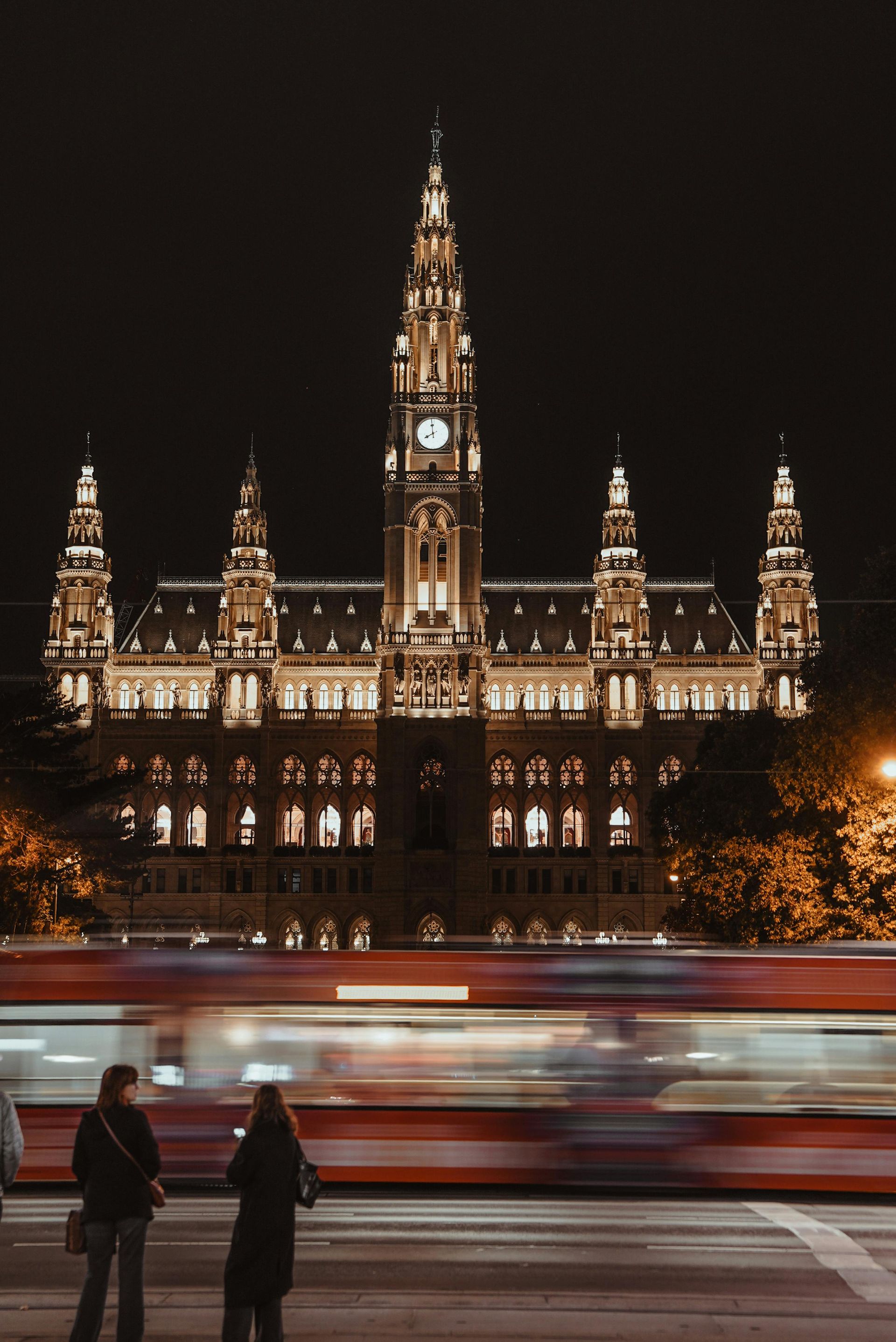 Lit-up historic city hall at night with blurred traffic passing in front.