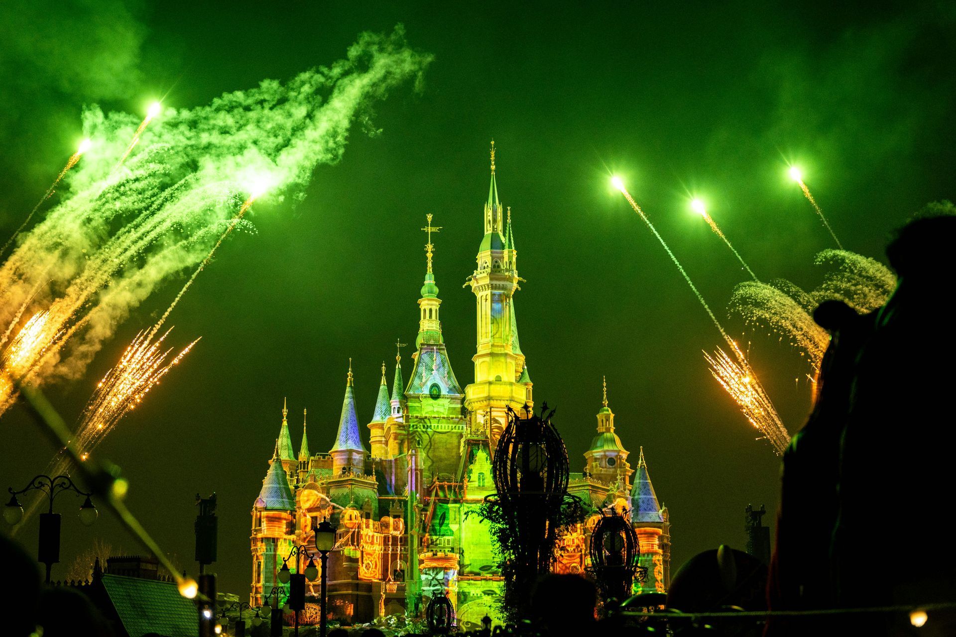 A brightly illuminated Disney castle at night, framed by vibrant green fireworks exploding in the dark sky.