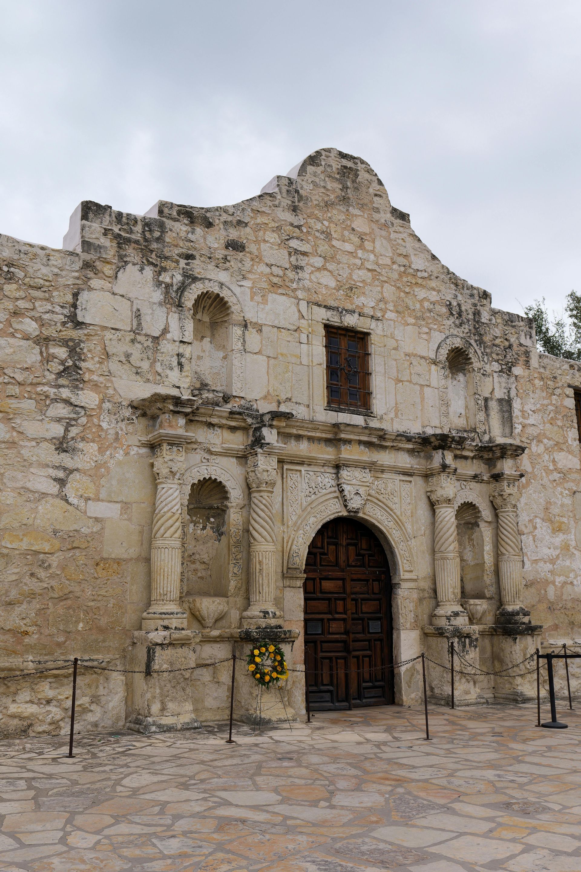The limestone facade of The Alamo mission in San Antonio, Texas, featuring a central arched wooden doorway and columns.