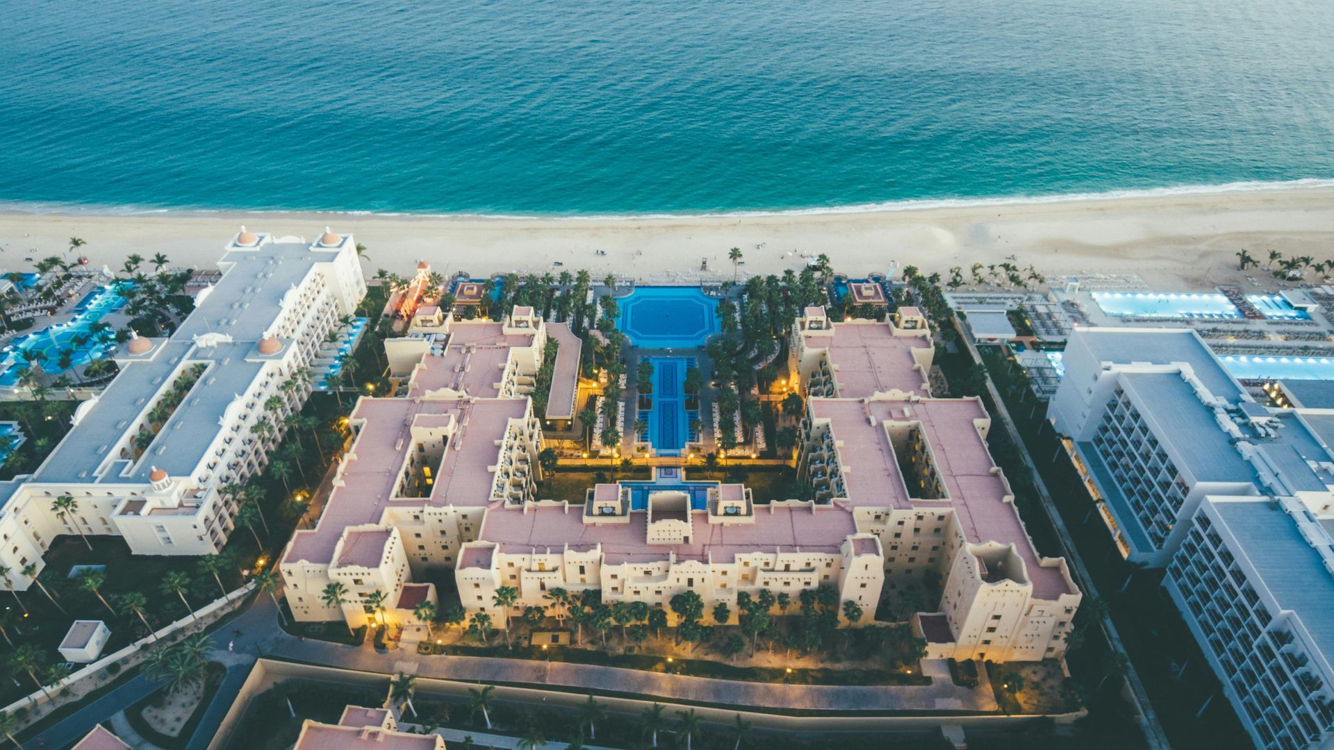 Aerial view of a beachfront resort with pink buildings, pools, and turquoise ocean beyond the sand.