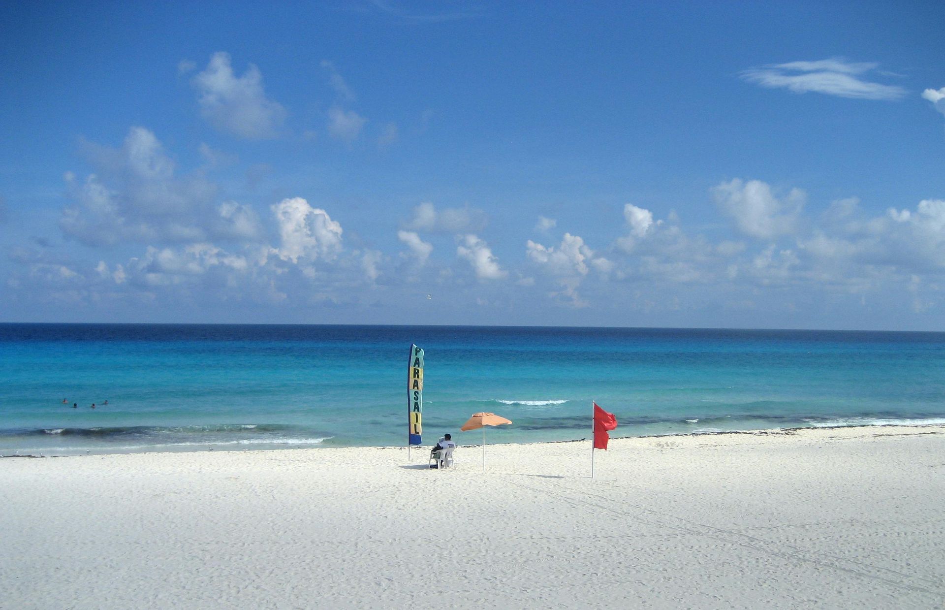 A person sits on white sand beach facing a turquoise sea under a bright blue sky with scattered clouds.