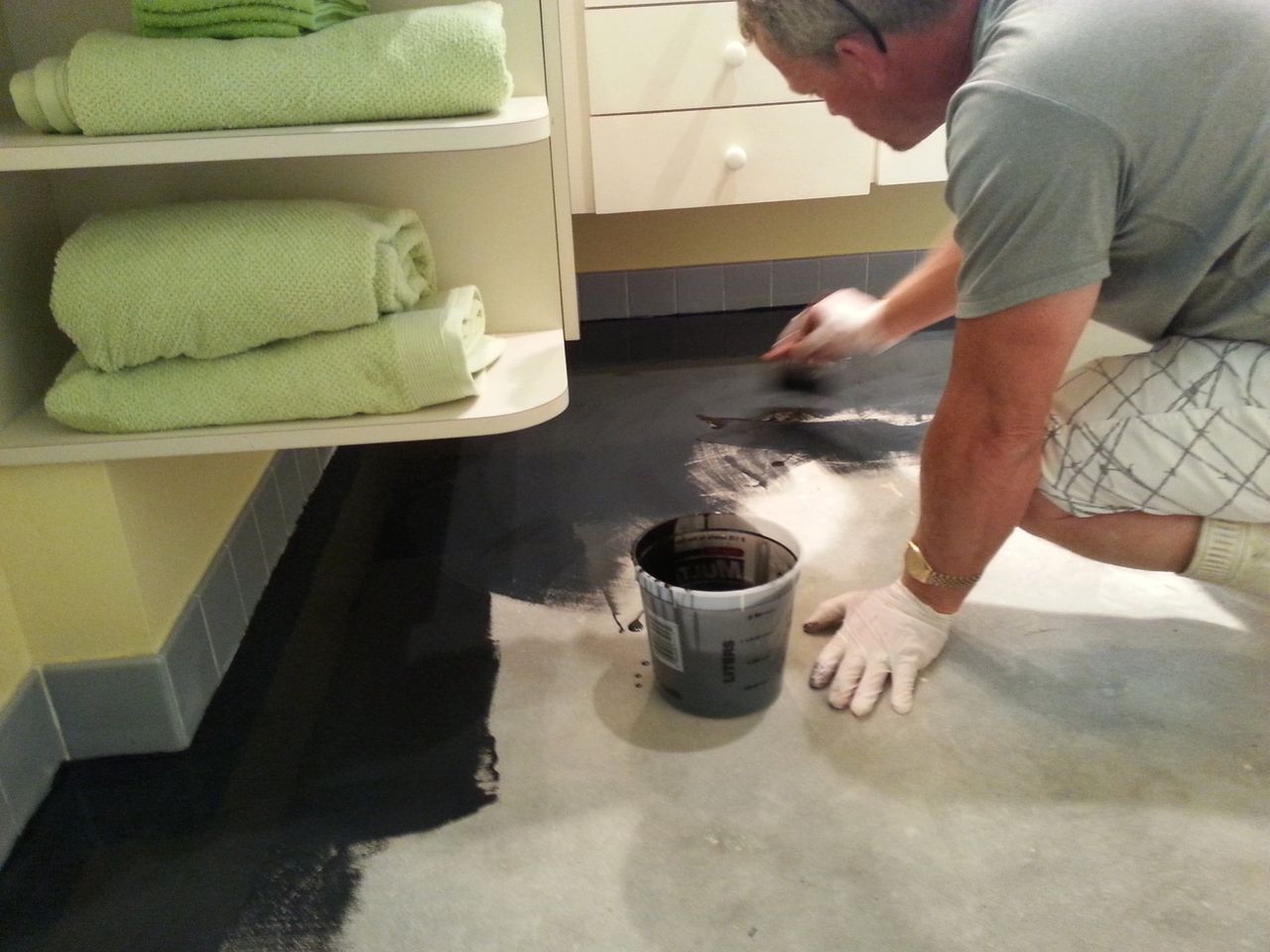 A person applies dark floor sealant to a light concrete floor in a room with light green towels on white shelving.