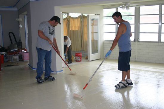 Two people use paint rollers to apply a light-colored coating to a residential floor.