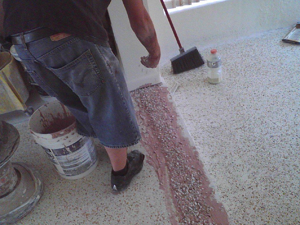 A person applying a reddish-brown textured sealant to a light-colored speckled concrete floor.