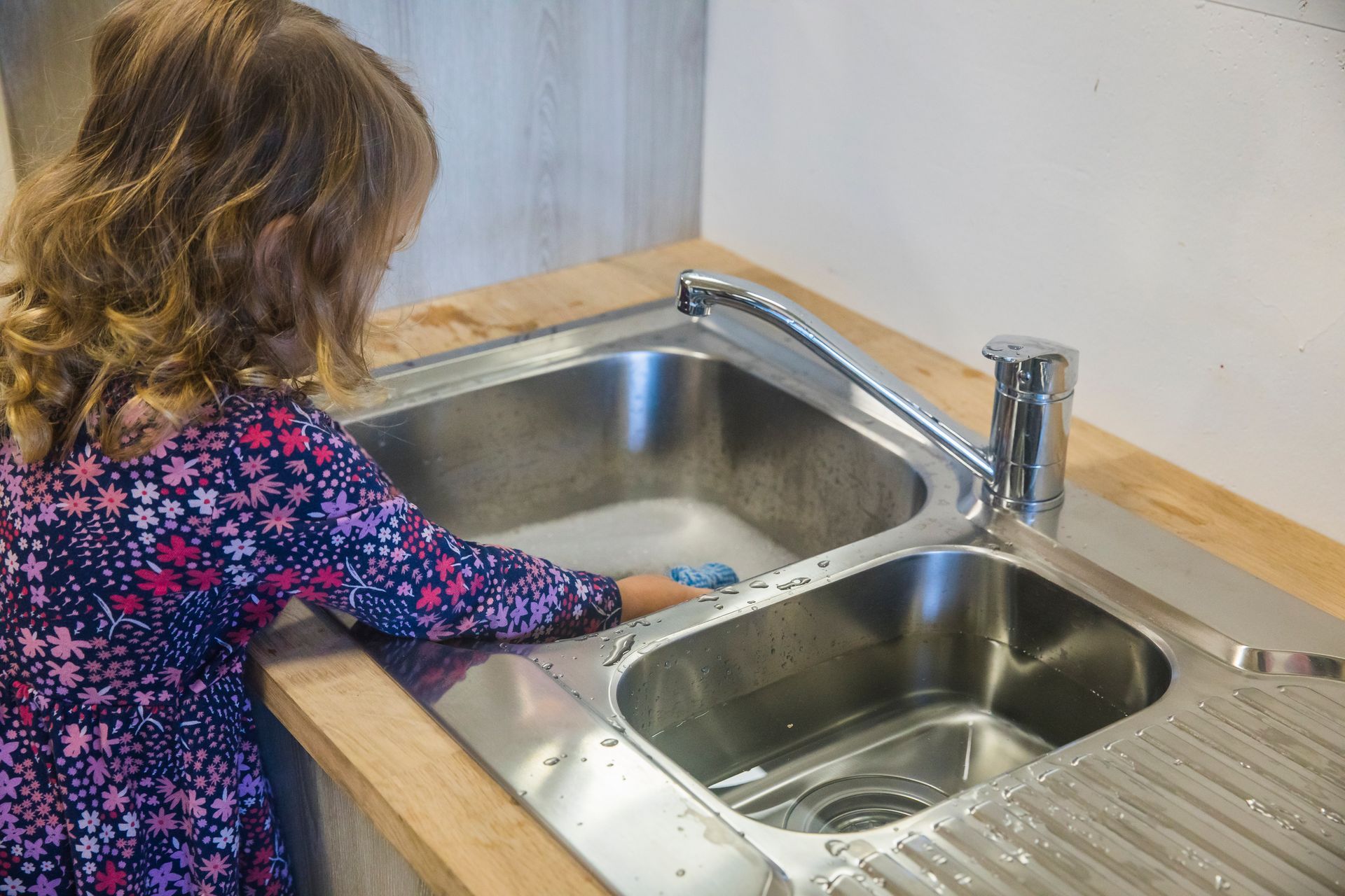 A little girl is washing dishes in a kitchen sink.