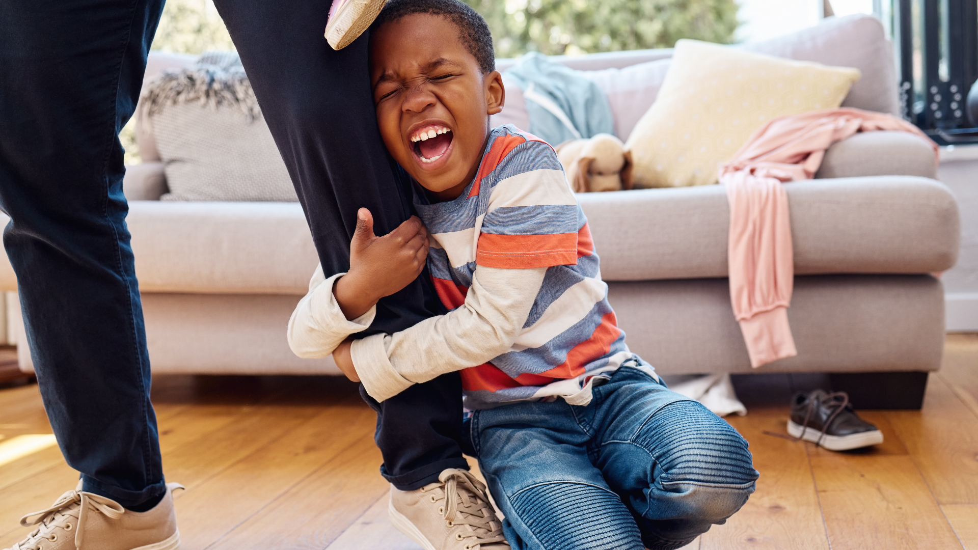 Boy crying, clinging to a leg, in a messy living room.